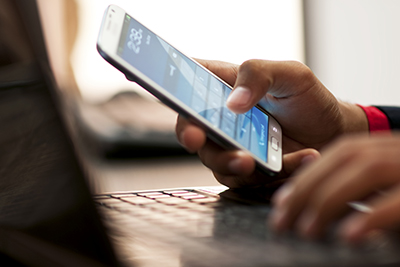 An image of a person looking at their cellphone and typing on their laptop computer.