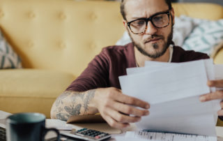 An image of a young man looking over paperwork in front of his laptop computer.