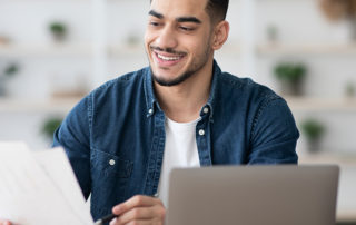 An image of a young man looking over paperwork at his laptop.